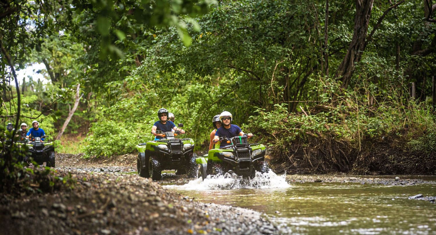 Multiple ATV riders splashing through a muddy puddle in a forested trail, illustrating Ontario’s dynamic off-road riding conditions.